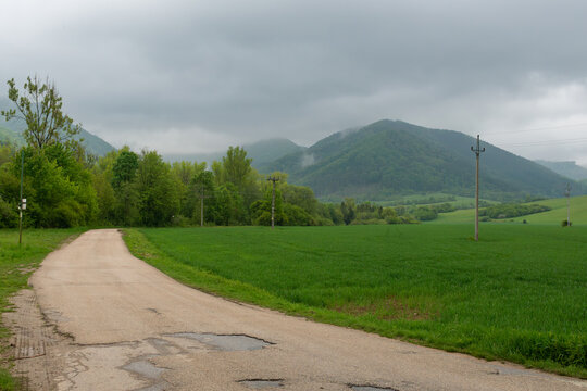 Quiet country road near Varin, Slovakia, running beside lush cereal fields toward forested mountains covered by low clouds and drifting mist. Utility poles, rolling farmland and dramatic rainy weather