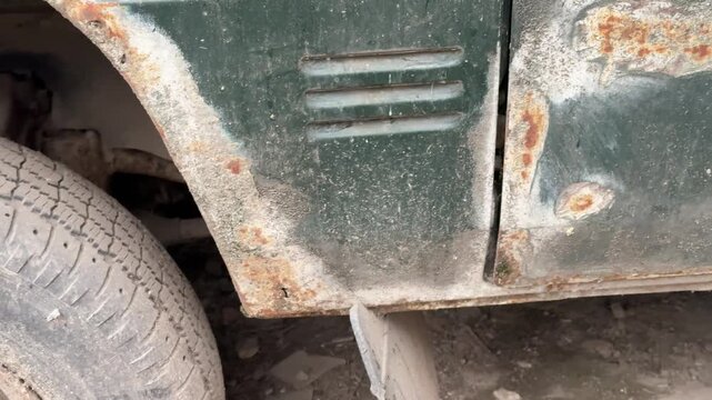 Abandoned dark green minibus with punctured tire, close-up of muddy wheel and spare rim tucked under chassis, heavy rust patches