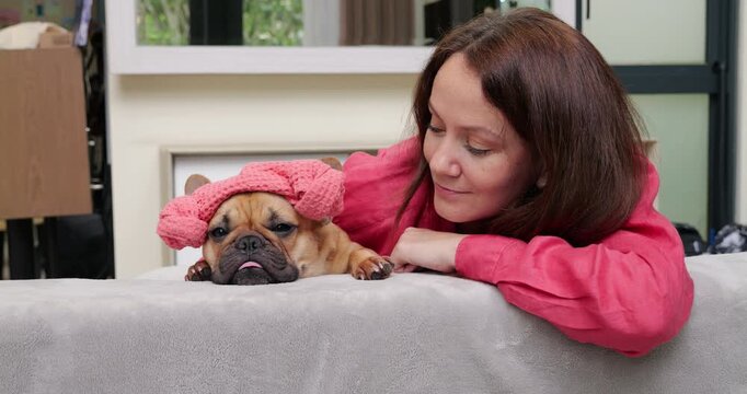 Adorable French Bulldog wearing pink spa hat resting peacefully beside long-haired woman in pink shirt, sharing cozy moment on home sofa. Portrait shot, both look to camera at first