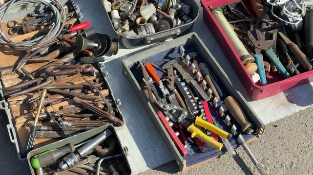 Tools for sale on asphalt showing assorted pliers, wrenches, screwdrivers and sockets in open boxes jars of screws and rusty
