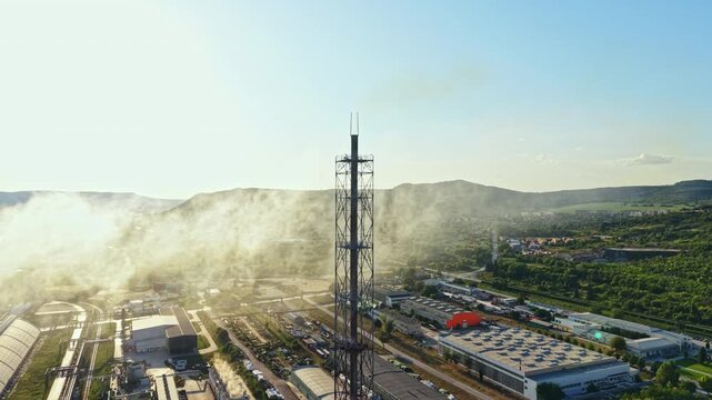 A tall telecommunication tower stands in a rural area surrounded by buildings and smoke. Hills rise in the background under a clear sky during late afternoon hours.
