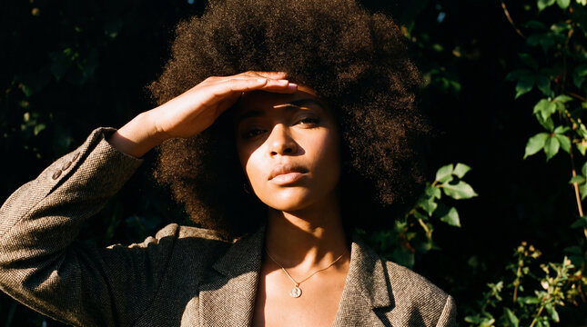 Portrait of a young Black woman with an afro shielding her eyes from sunlight outdoors against green foliage