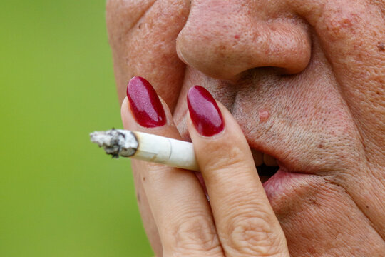 Close-up of hand with red nails holding cigarette. Smoke curls from cigarette tip. Face partially visible, showing wrinkles and skin texture.