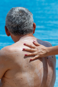 Man applies sunscreen to his back near a pool. Woman's hand with red nails touches his skin.