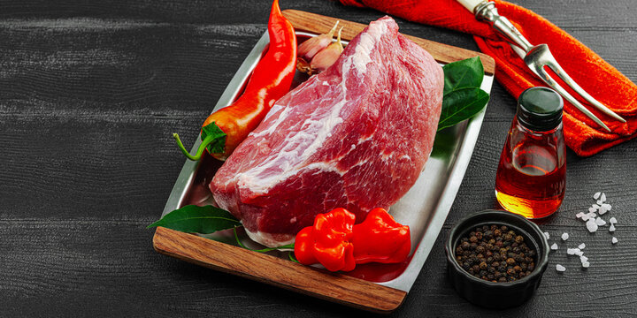 Raw pork loin on a metal tray with chili peppers, garlic, bay leaves, oil, and spices