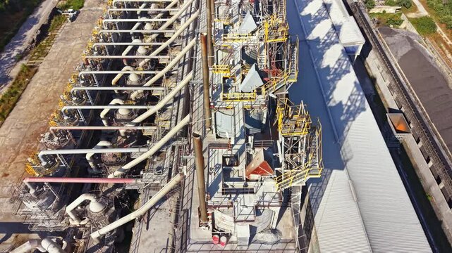 Heavy machinery works at a large industrial facility. Storage silos stand tall in rows. The setting is outdoors with clear skies. Workers move around the site engaged in tasks.