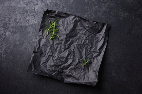 Black parchment paper with rosemary sprigs on a dark textured background. Close up. Copy space. Top view.