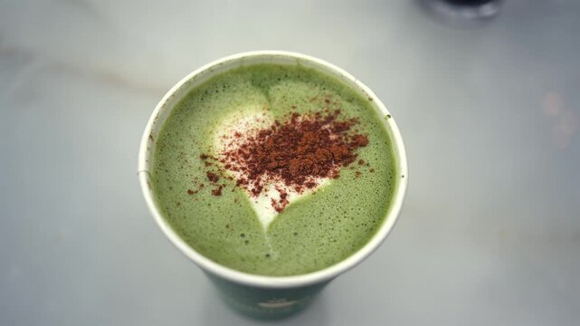 Matcha green tea latte in paper cup with heart shape foam and cocoa powder topping cinematic motion.