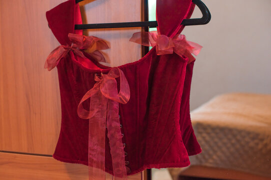Luxurious deep red velvet corset top with off-the-shoulder straps and ribbon bows hanging on a hanger or lying in a gift box. Elegant feminine fashion lingerie garment with lacing detail