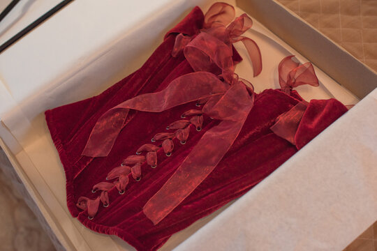 Luxurious deep red velvet corset top with off-the-shoulder straps and ribbon bows hanging on a hanger or lying in a gift box. Elegant feminine fashion lingerie garment with lacing detail