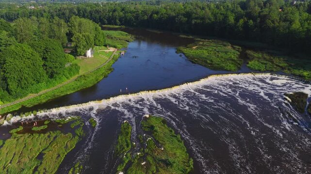 Aerial view shows Venta Rapid at Kuldiga, Latvia, a wide low waterfall across the Venta River, with grassy islets, promenade, small white building, buoys, and people wading.