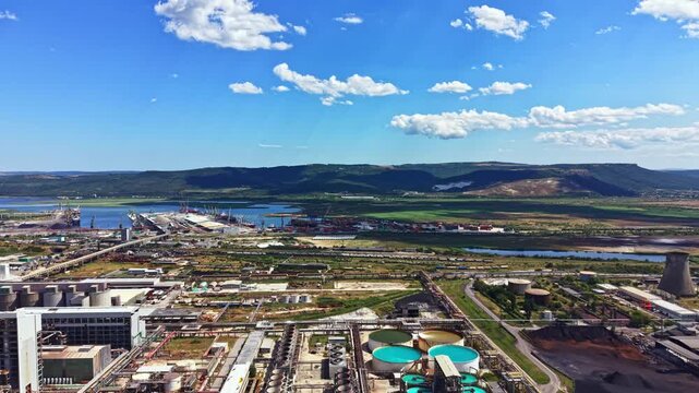 The view shows a vast industrial area with factories, large water tanks, and a river. Rolling hills and open fields are visible in the distance under a blue sky with clouds.