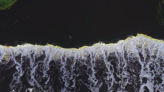 Aerial top down view of Venta River at Kuldiga, Latvia. Venta Rapid forms a long low crest with yellow tinged foam, dark water, swirling motion, and a tiny human.