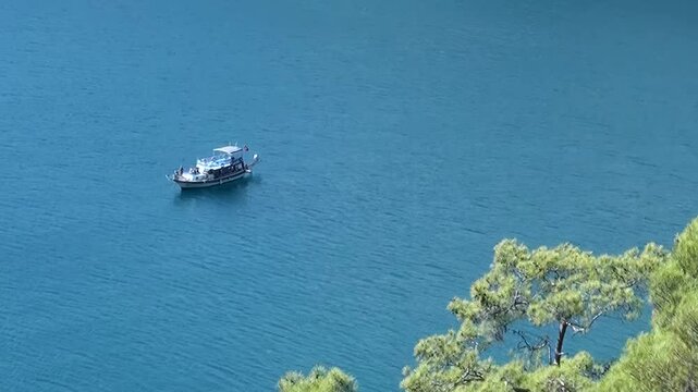 Overhead view captures a boat peacefully drifting on the dazzling azure surface of Oludeniz Bay, Turkey. Sunlight dances on the calm water beside verdant Mediterranean hills.