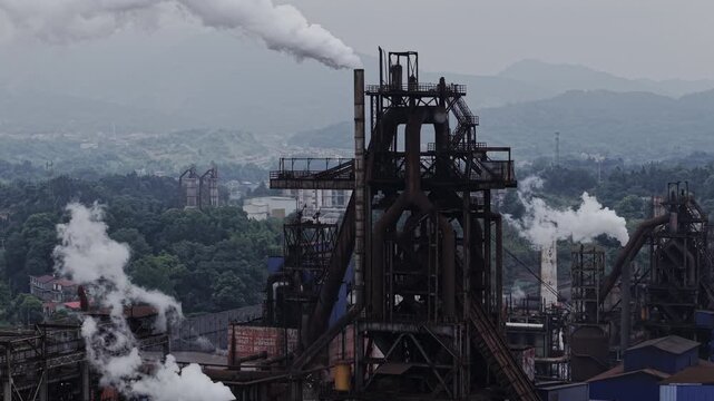 Drone slowly ascending above an industrial factory with heavy steam rising continuously from chimneys against dark mountains. Aerial shot of carbon emissions.