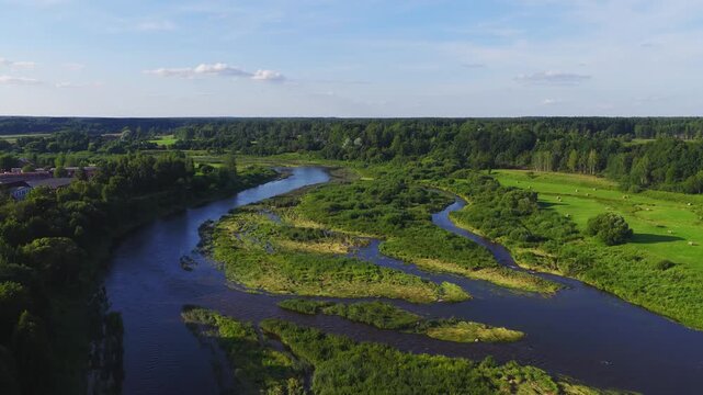 Aerial drone pans above the Venta River near Kuldiga, Latvia, showing sinuous channels, islands, hayfields, woodland, hay bales, and marshy reeds on a sunny afternoon.