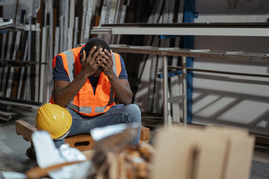 A burnout  factory worker sits on a pallet holding his head in his hands after a long and stressful shift in an industrial workplace.