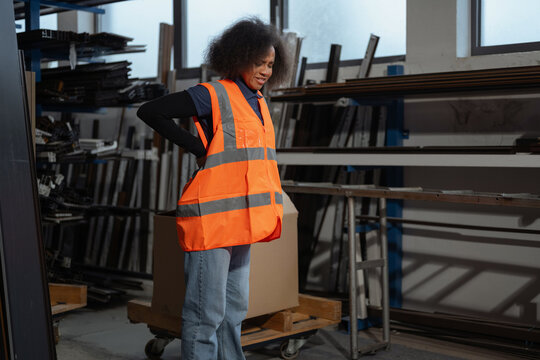 A woman wears safety gear and holds her back in a factory while lifting a heavy cardboard box. This shows risks of manual handling.