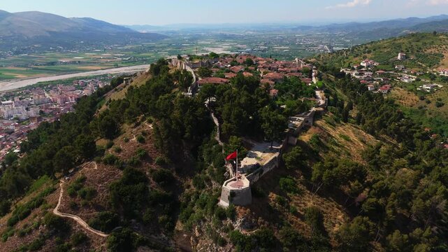 Aerial drone footage approaching Berat Castle on its wooded hilltop, showing the historic settlement within the fortress walls, traditional red-tiled roofs and the broad Albanian valley beyond.