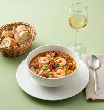 A bowl of hot white bean soup with shrimp, sausage, and bacon, accompanied by a basket of rustic bread and a glass of white wine.
