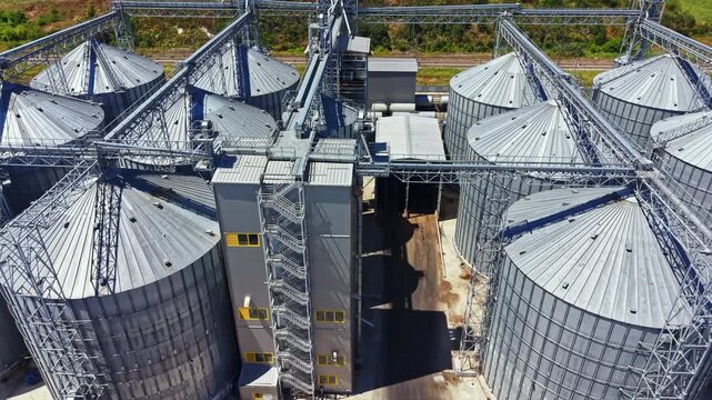 A large storage facility with multiple grain silos sits in a rural area. The silos are connected by metal structures. This view shows the layout of the facility during daylight.