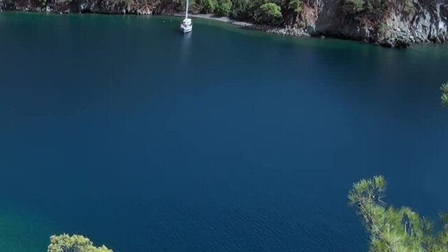 Elevated view reveals Fethiye Turkey's stunning coastline on bright day. Graceful white sailboat floats peacefully on deep blue Mediterranean Sea, nestled against verdant cliffs and lush green trees.