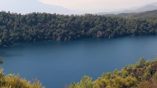Discover the serene splendor of Oludeniz Bay, Turkey. Azure waters gently caress verdant forested hills, while a majestic mountain range fades into the distant horizon under a bright, peaceful sky.
