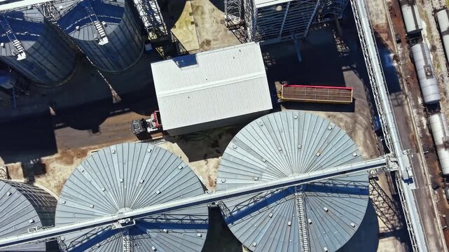 Bird's eye view shows large round storage tanks at a grain facility. The tanks are arranged in rows. Metal structures connect them. Trucks are present on site. The setting is active and busy.