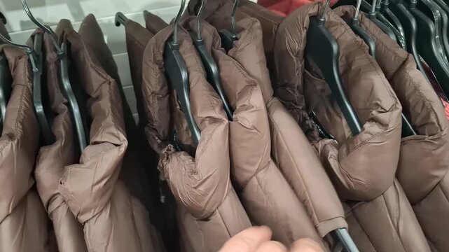 A man's hand chooses a brown puffer jacket from a clothing rack in a retail store, shopping for winter outerwear, winter clothes shopping, retail store interior.
