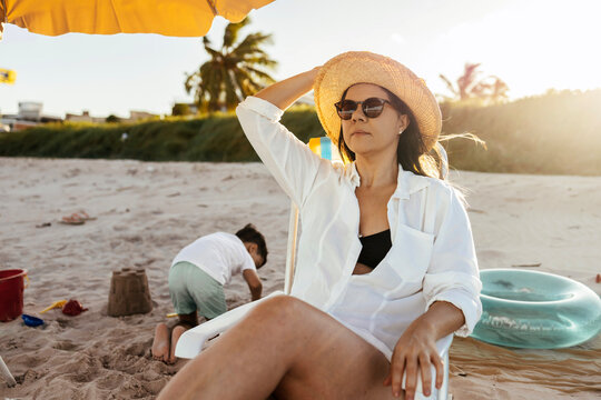 A mother relaxes in a beach chair under an umbrella as her young son concentrates on building a sandcastle nearby. Warm sunlight, seaside breeze and family vacation vibes create a peaceful scene.