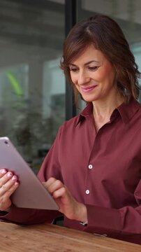Middle aged businesswoman using digital tablet in office. Smiling mature woman financial advisor holds device, checks business data online. Female expert consults clients at desk. Vertical
