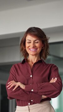 Portrait of confident businesswoman in 40s standing with arms crossed in office. Mature woman boss looks at camera with light smile. Trustworthy female business chief leads company. Vertical