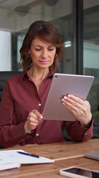 Working on digital tablet in office, latin mature businesswoman reading report online. Focused woman startup founder using device at desk. Female owner grows business company at workplace. Vertical