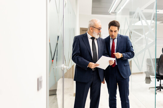 Two people between 40 and 60 years old, wearing suits, are talking in the middle of a hallway with papers in a modern office. Concept of lawyers or businesspeople.