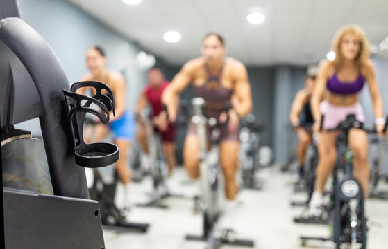 A group of people are exercising on stationary bikes in a gym. In the foreground is a water bottle holder. Concept of stationary bike classes.