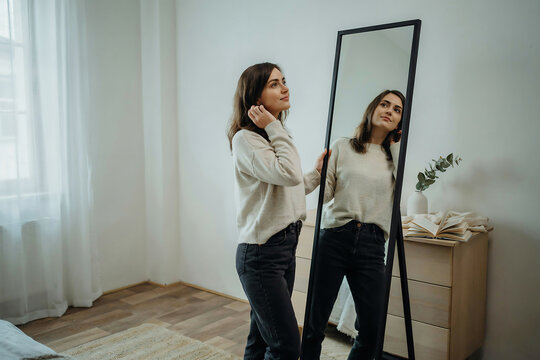 Woman in beige sweater and black jeans looking at her reflection in a full-length mirror