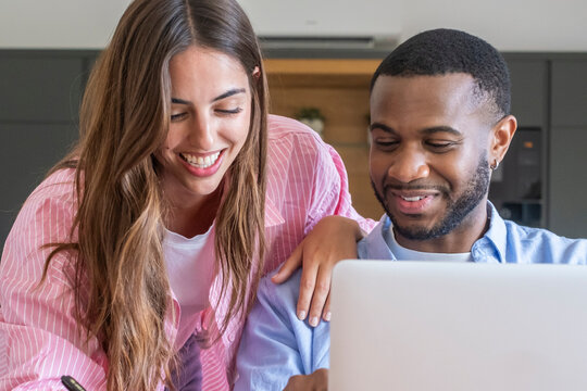 Multicultural couple laughing and enjoying collaborative work on a laptop, representing teamwork, happiness, and technology in a modern home office setting