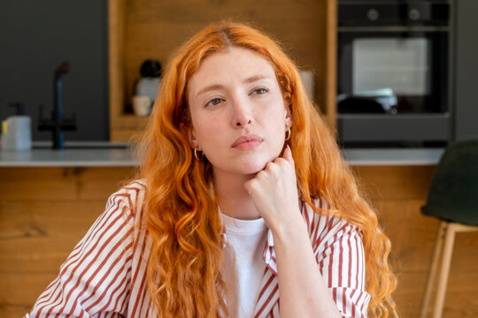 Redhead woman with wavy hair looking away and resting her chin on her hand, contemplating in a modern kitchen setting