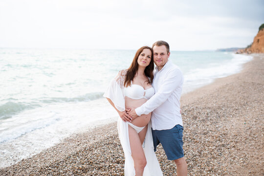 Smiling pregnant woman wearing bikini walking at beach with happy man holding belly over sea. Maternity clothes.