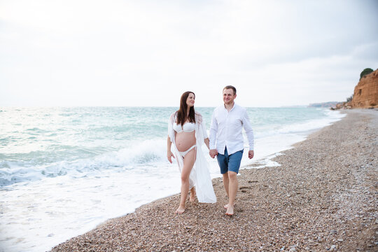 Smiling pregnant woman wearing bikini walking at beach with happy man over sea. Maternity clothes.