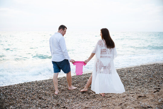 Pregnant woman wearing bikini at beach with happy man holding baby onesie over sea. Maternity clothes.