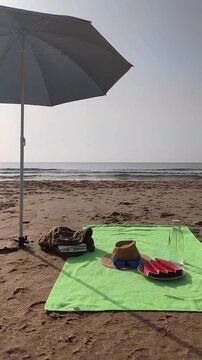 Vertical video of a beach towel spread on golden sand with a book, a plate of freshly cut watermelon, and a bottle of fresh water resting on it, while the sea moves gently in the background
