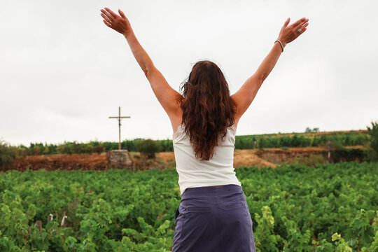 A woman raises her arms toward a cross overlooking vineyards in the Burgundy wine region of France, expressing joy and connection with the peaceful landscape.