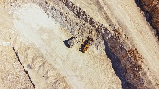 Two large machines operate in a quarry. They move earth and rocks while extracting materials. The site shows signs of recent activity and change. Dust rises from the ground.
