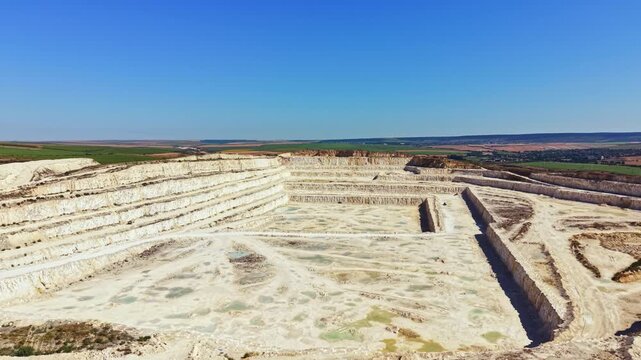 A large quarry shows layers of stone and earth. The site is situated in a rural location with fields in the distance. Bright sunlight illuminates the structures and landscape.