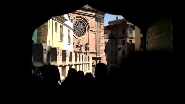 Sunlit historic street framed by dark archway, ornate church facade, circular rose window, pastel buildings, tiled rooftops, silhouetted pedestrians, layered urban depth, warm stone textures, clear