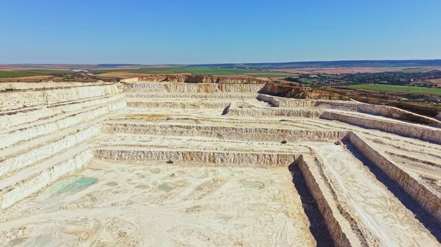 Deep quarry shows large sections of rock that have been removed. Machines work in the distance while the landscape reveals layers of earth. Clear sky allows for bright daylight.