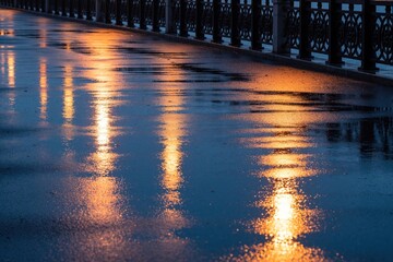 Fototapeta premium Bright orange street lamps reflect on wet dark asphalt pavement after rain at night. Metal railing lines walkway along city embankment path. Gloomy urban scenery creates moody atmosphere with golden