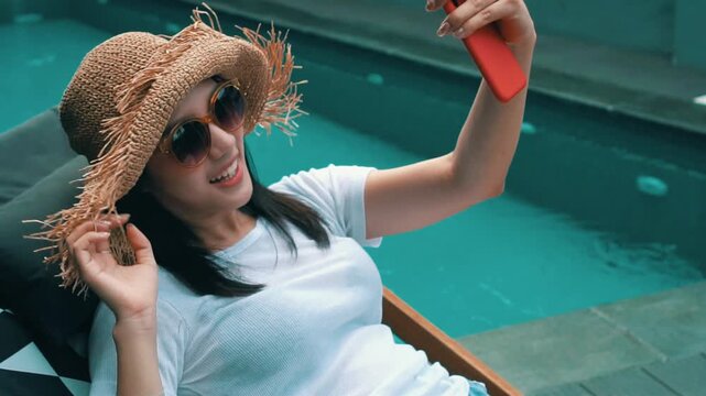 Happy young Asian woman in straw hat and sunglasses taking selfie or video calling on smartphone while relaxing on sunbed near swimming pool. Cheerful female enjoy summer vacation and tropical resort