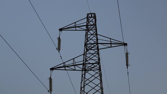 Close-up shot of the upper section of a high voltage electric pylon with power lines and insulators against a plain sky background.